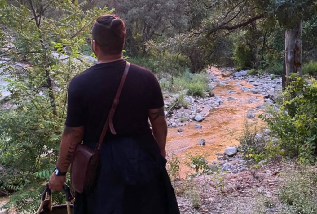a man with a basket slung over a shoulder looks into the woods