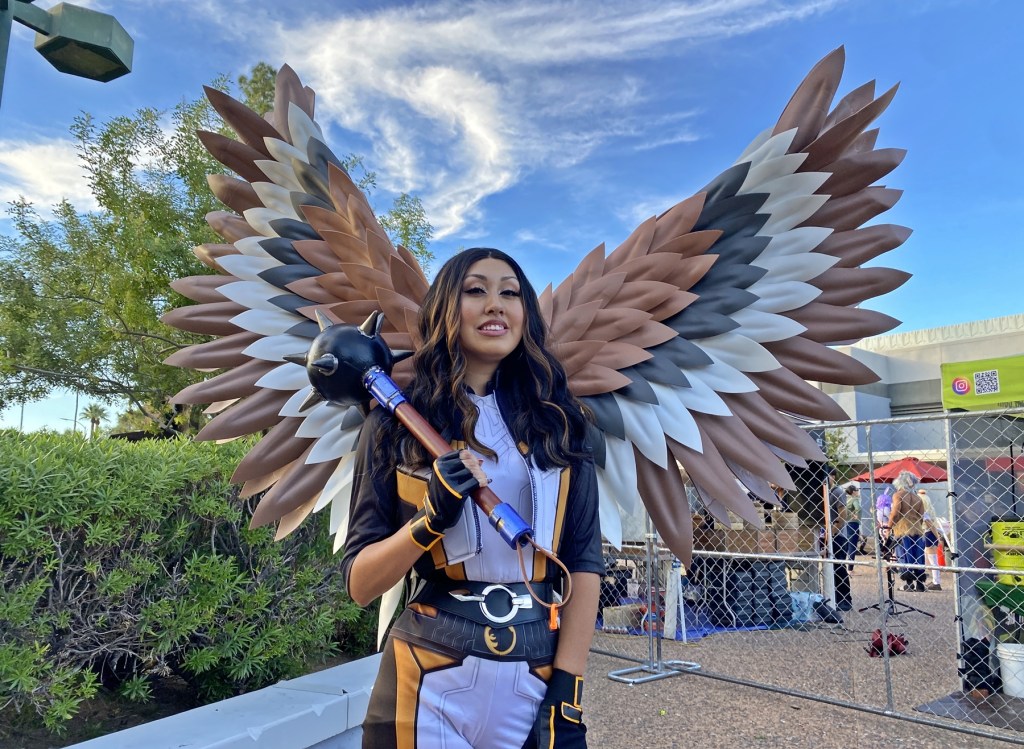 A female cosplayer dressed in a winger superhero costume stands outside of a convention center.