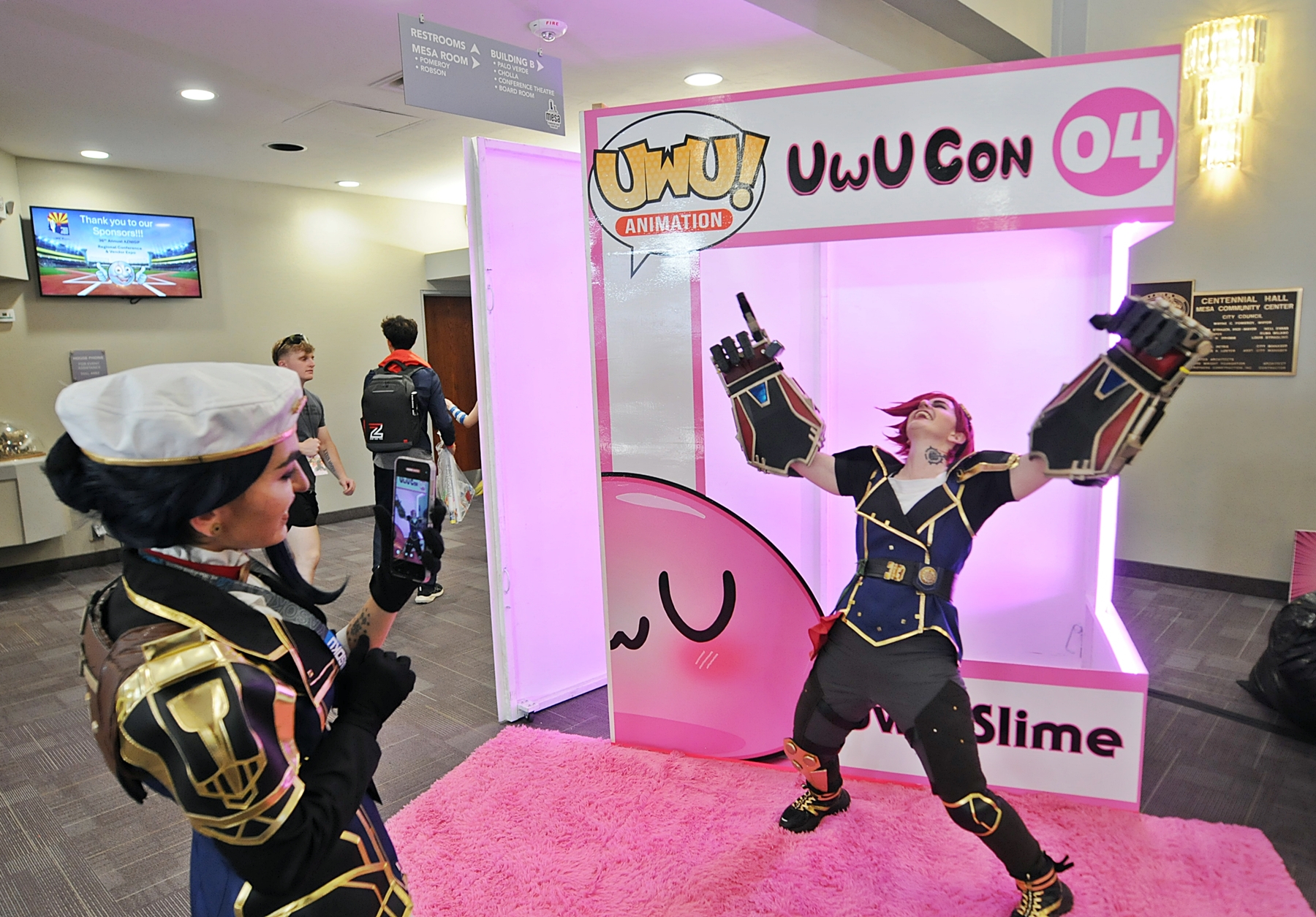 A cosplayer takes a photo of her friend in costume in front of a giant pink display shaped like a box.