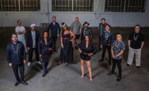 A group of 13 musicians standing in front of a gray wall.