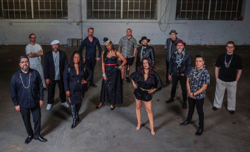 A group of 13 musicians standing in front of a gray wall.