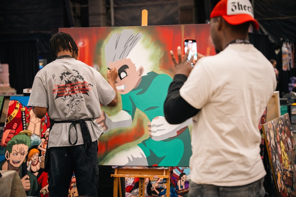 A male takes a photo of an artist creating an anime painting.