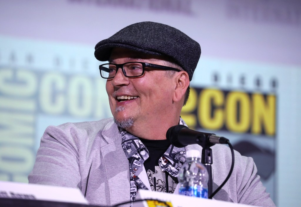 An older man in glasses and a hat smiling behind a microphone.
