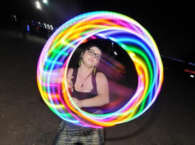 A young woman in a black tank top holds a rainbow-colored LED hula hoop at night.