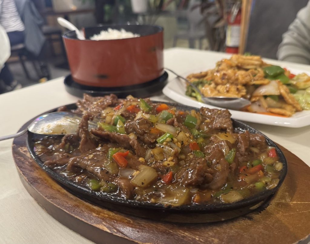 A table with a sizzling plate in the forground, filled with other Chinese food dishes.