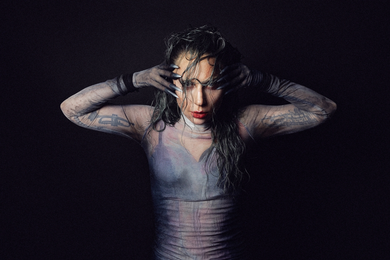 A long-haired woman in a mesh top and bra looks directly at the camera.