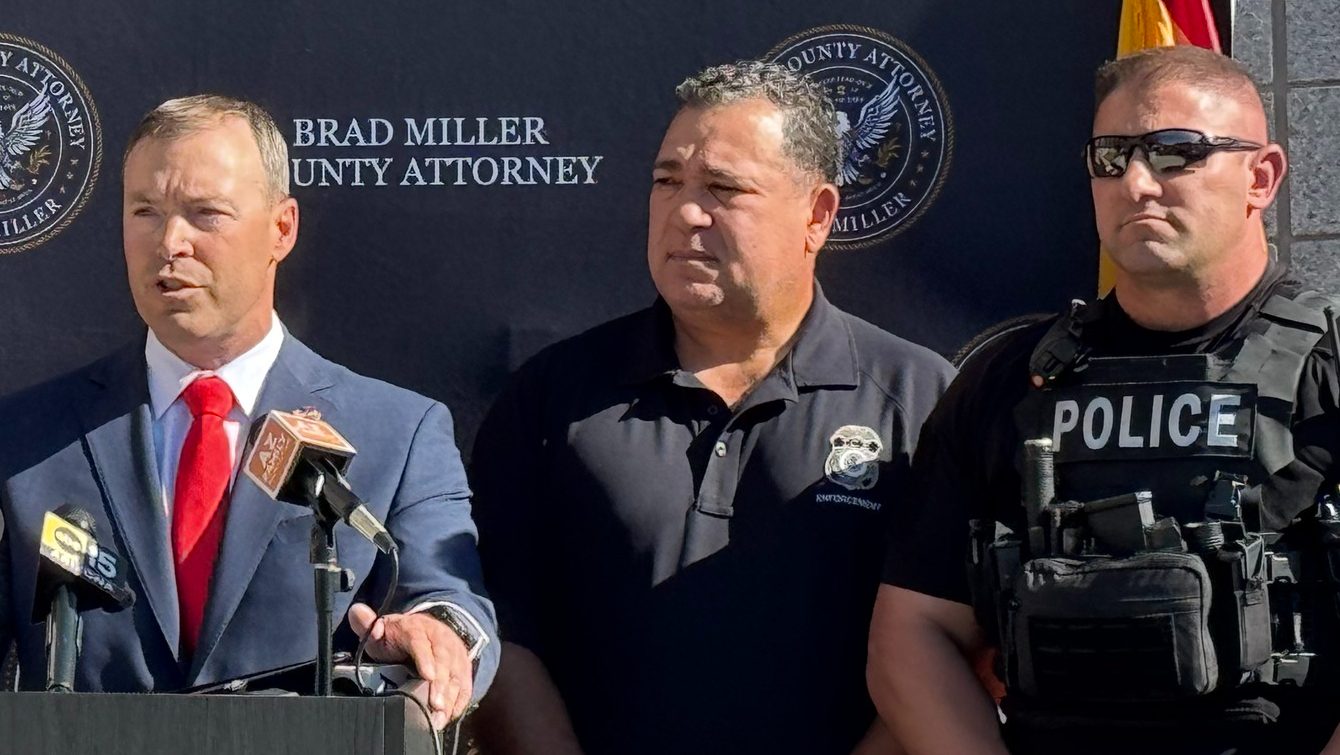 a man in a suit speaks at a podium, flanked by two other men -- one in a polo and another in a police vest