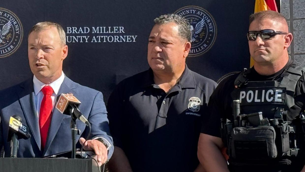 a man in a suit speaks at a podium, flanked by two other men -- one in a polo and another in a police vest
