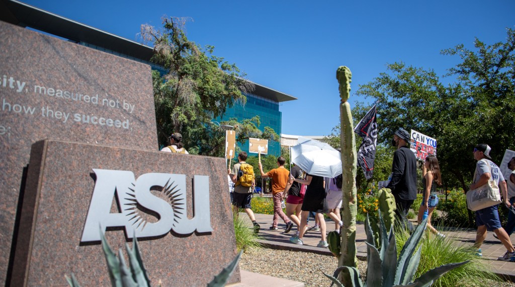 protesters march past an monument with the ASU logo