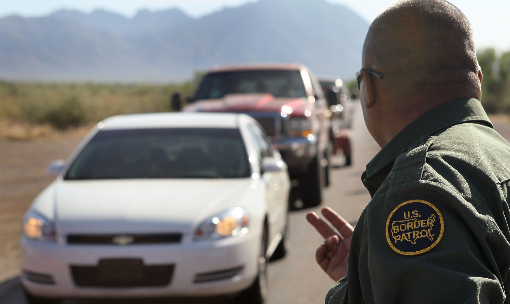 a border patrol agent waves forward a line of cars in a desert landscape