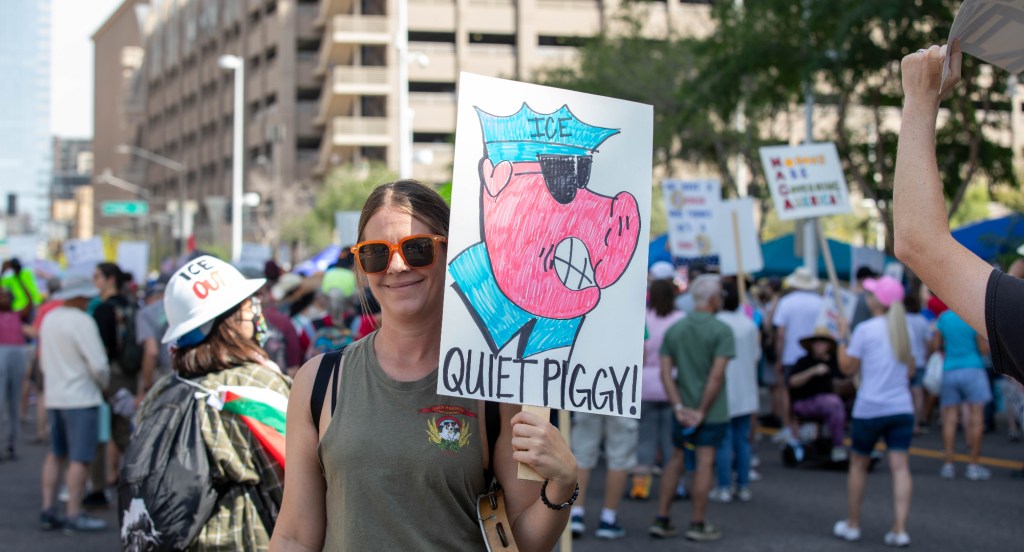 a protester holds a sign with an illustration of a pig in an ICE uniform, with the caption "quiet piggy"