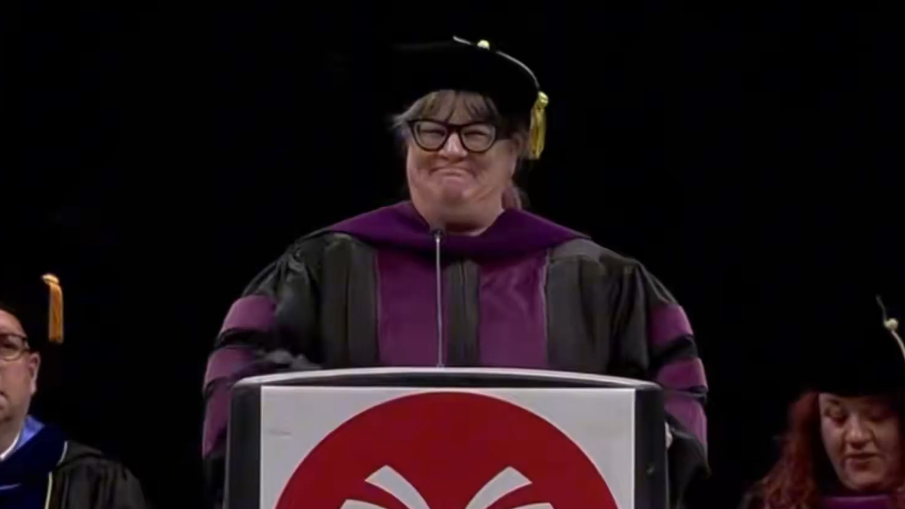 a woman in graduation garb stands at a podium with an awkward smile on her face