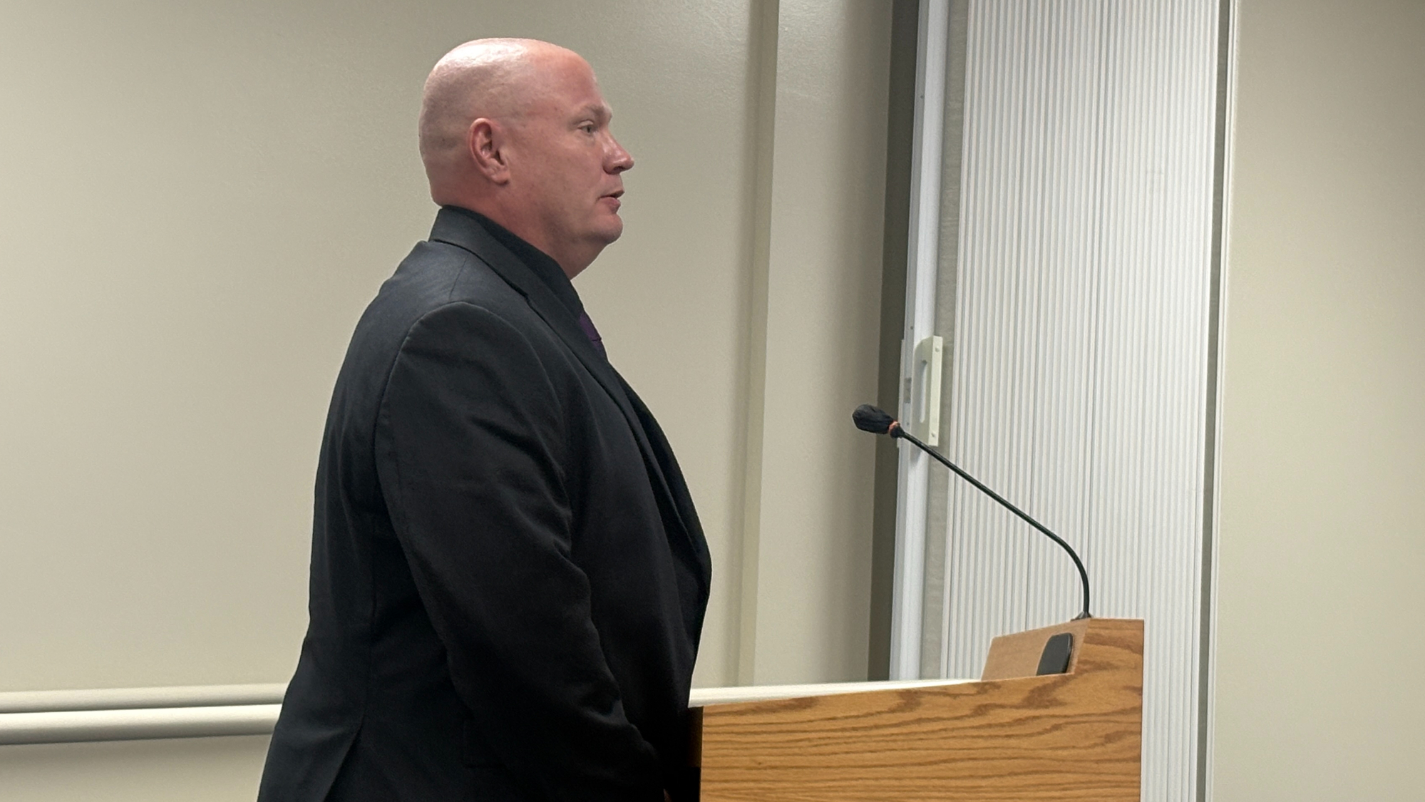 chuck trendle stands at a podium in a black suit