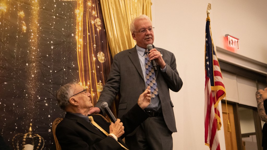 john kavanagh stands and speaks into a microphone next to a seated joe arpaio