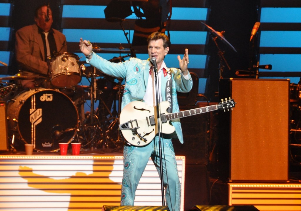 Chris Isaak performs onstage with a white guitar during a concert. The singer-songwriter is scheduled to play Celebrity Theatre in Phoenix on Oct. 13, 2026.