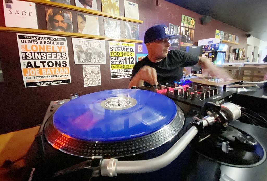 DJ Blesd1 performs at turntables inside The Record Gallery, a Phoenix record store lined with vinyl records and music posters.