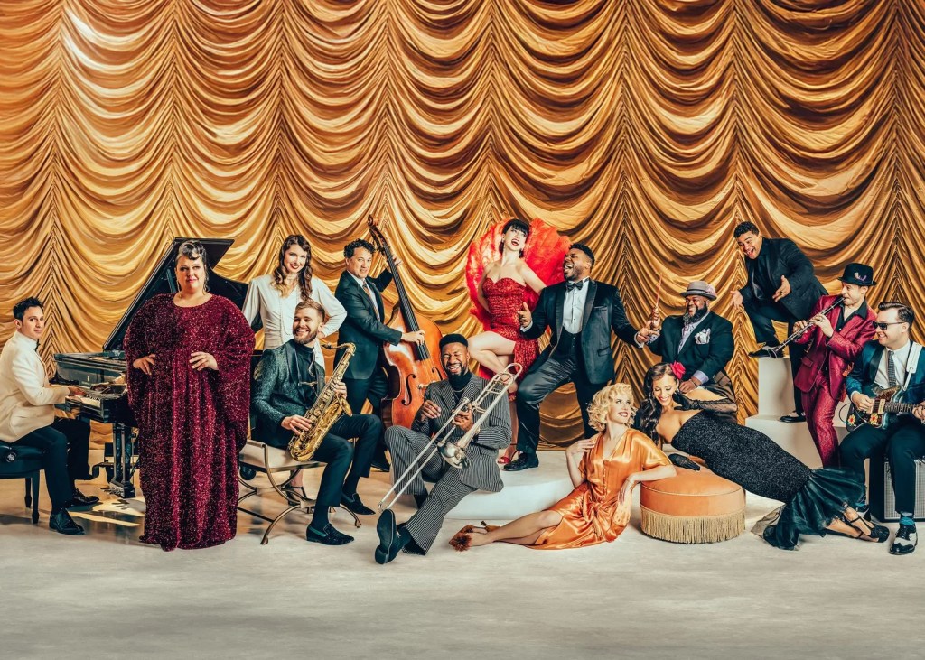 Members of Postmodern Jukebox pose in vintage-inspired attire in front of a gold stage curtain.