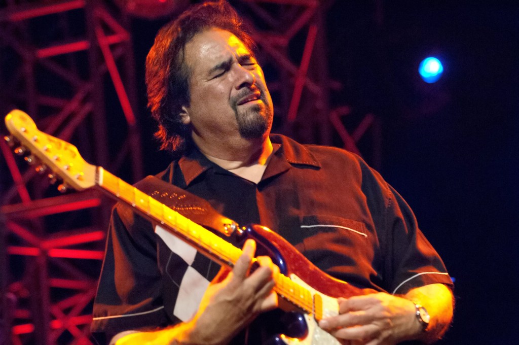 A middle-aged bearded man plays an electric guitar with his eyes closed.