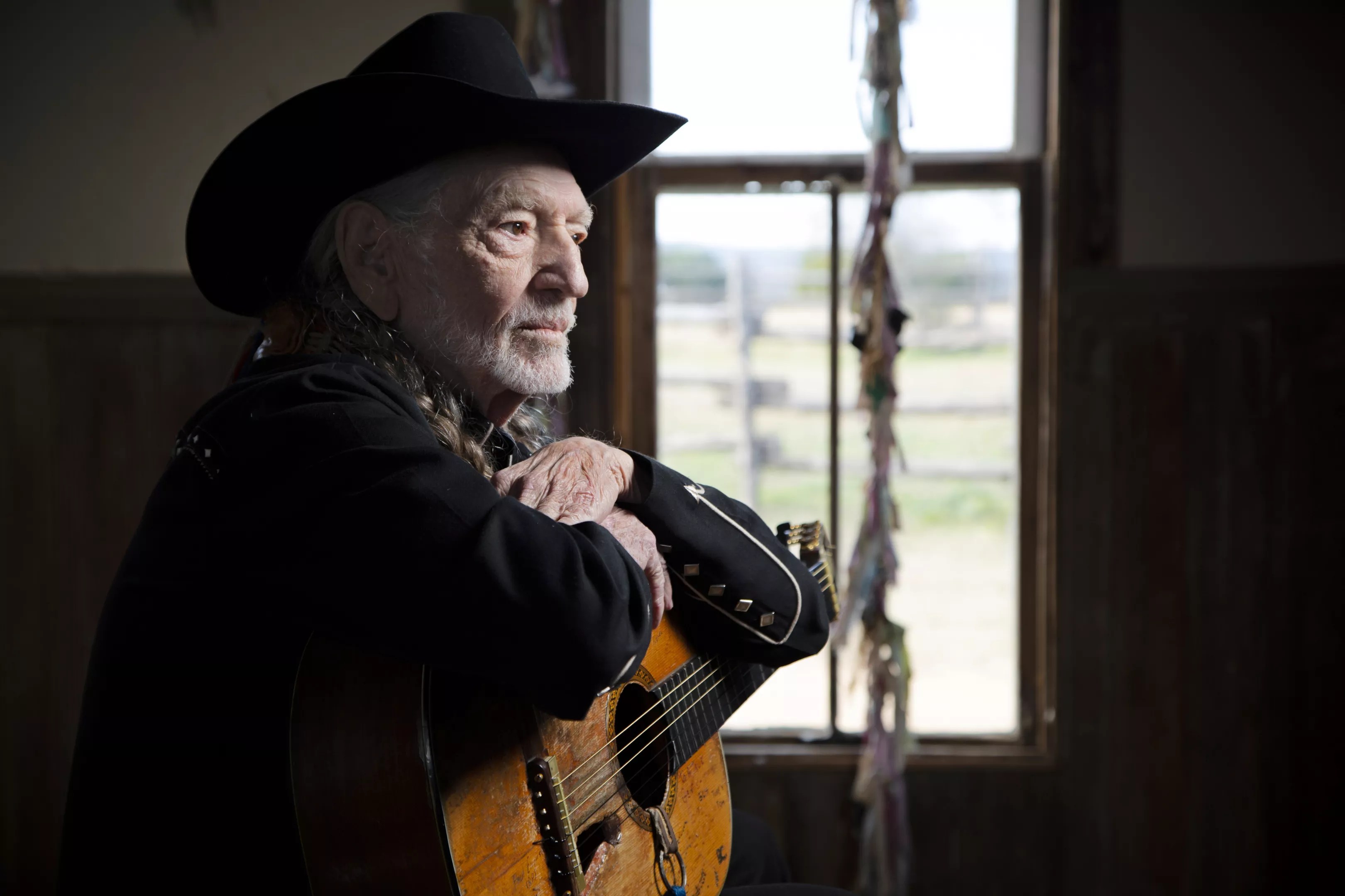 Willie Nelson sits in a room holding a guitar.