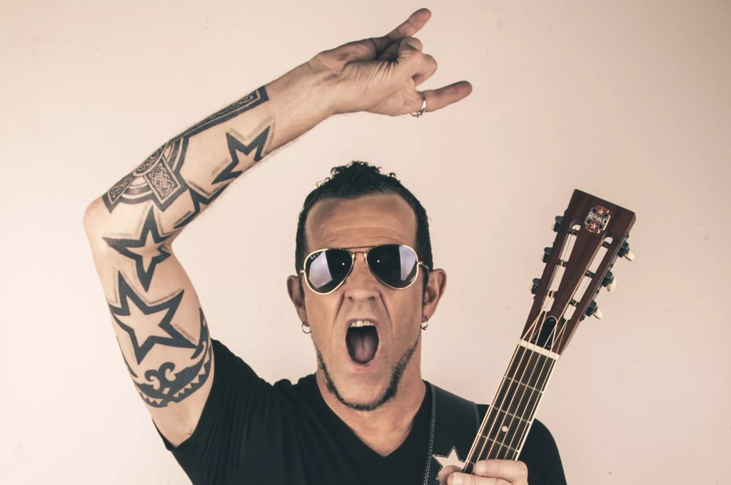 Tattooed rocker wearing sunglasses and holding a guitar in one hand and doing devil horns with the other hand.