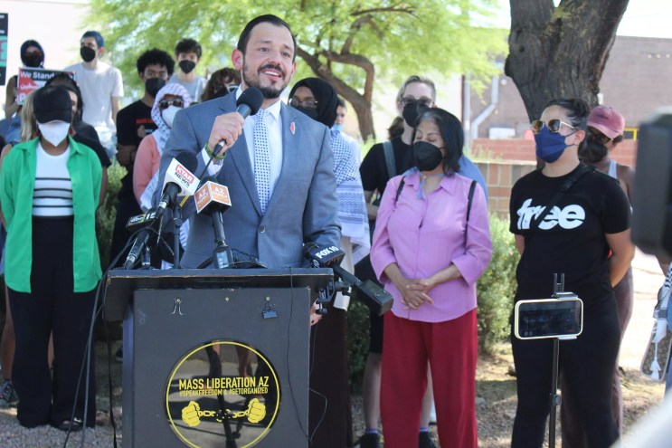 A man in a suit stands before a podium with a sign for "Mass Liberation AZ" and speaks into TV microphones. People in face masks stand behind him.