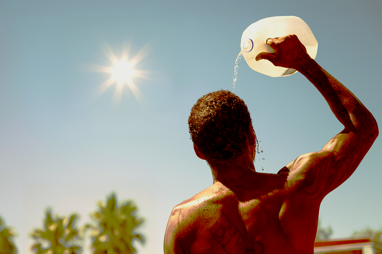 A man under a glaring sun pours water over his head out of a jug.