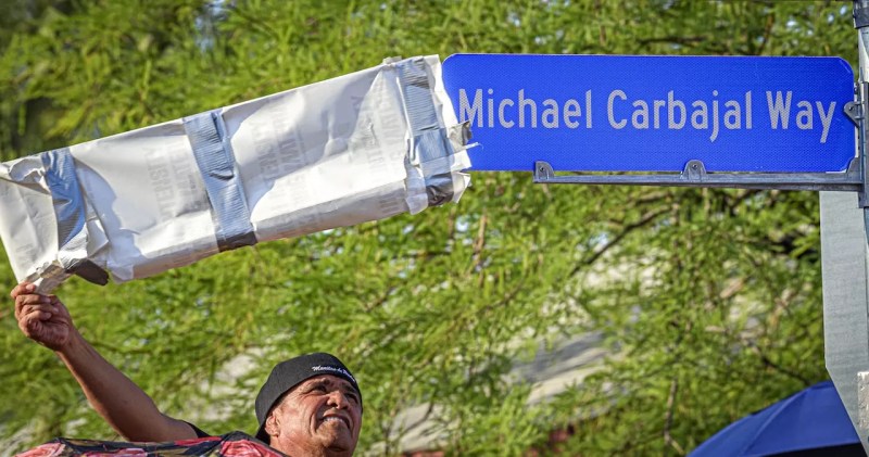 A man pulling a cover of a street sign that reads "Michael Carbajal Way"
