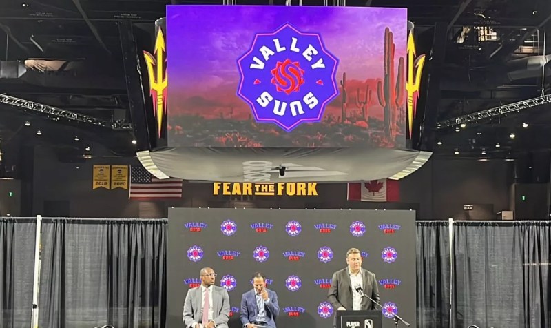 A man speaking at a podium on a stage. Above him is a video board showing the logo for the Valley Suns of the G League.