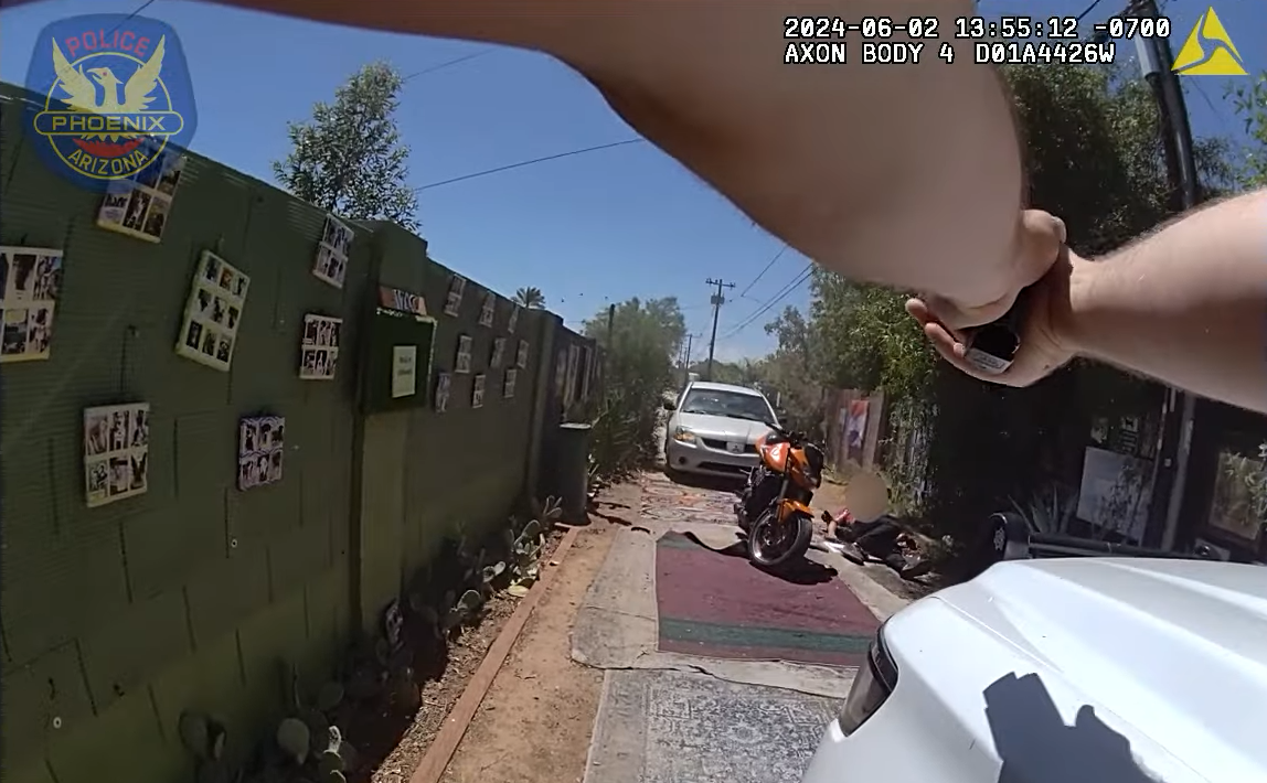 Body-cam footage showing two arms holding a gun on a blurred-out person who is laying on the ground next to a motorcycle.