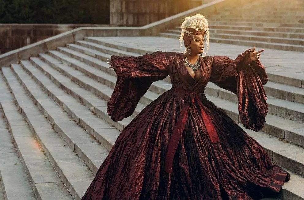A drag queen in a ballgown on the steps of a palace.
