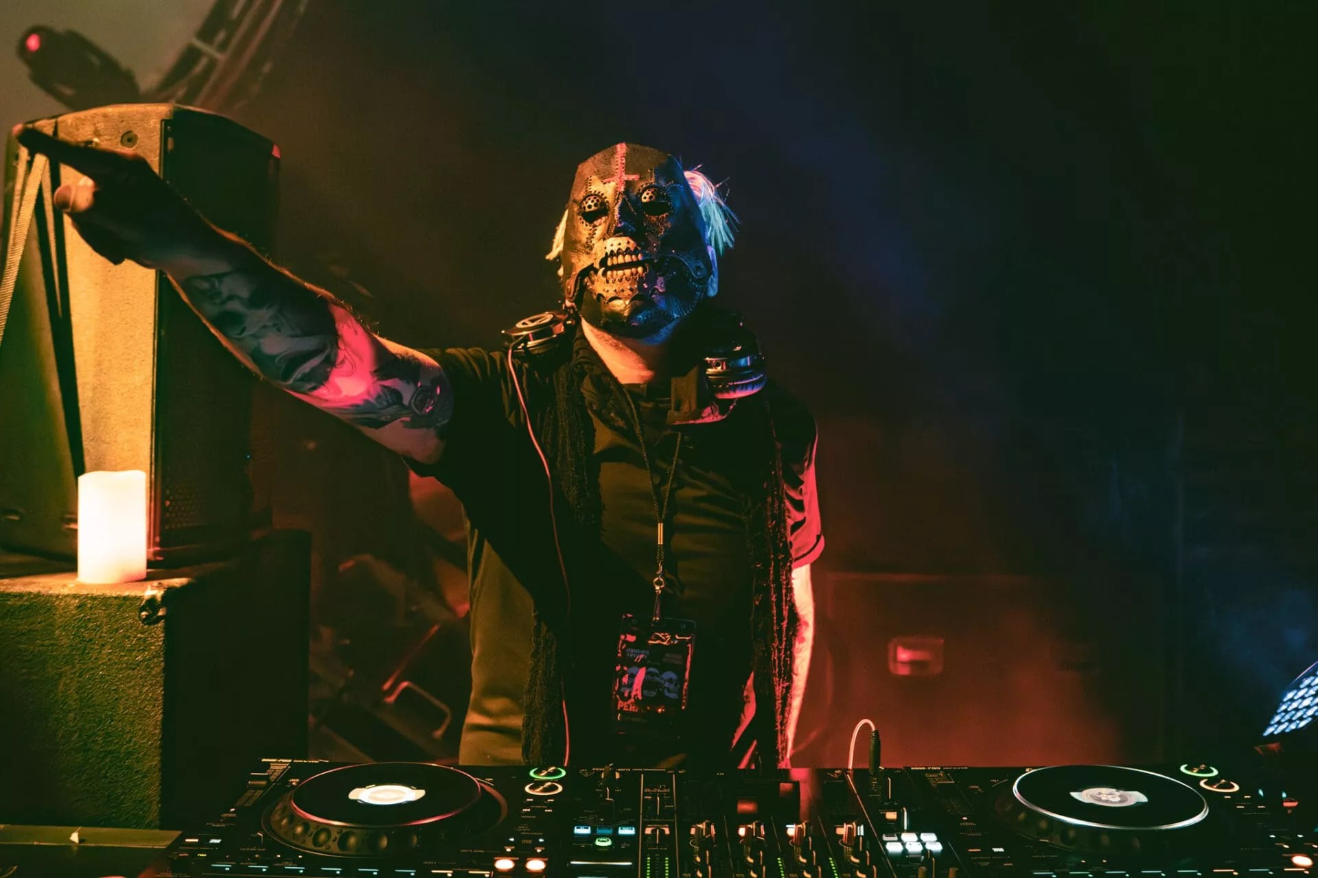 A masked DJ points from behind a mixer.