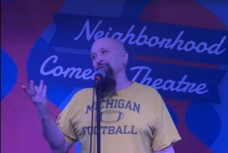 a man in a michigan football shirt on stage at a comedy club