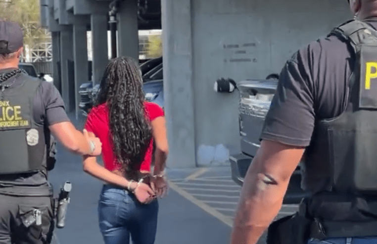 seen from behind, two police officers escort a handcuffed young black girl with braids