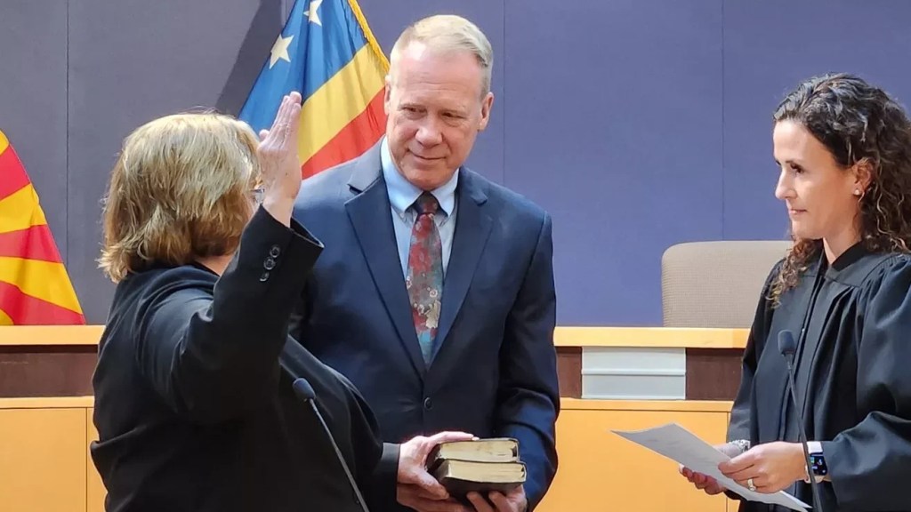 A woman raising her right hand with her left on a bible in front of a female judge. A gray-haired man holds the bible