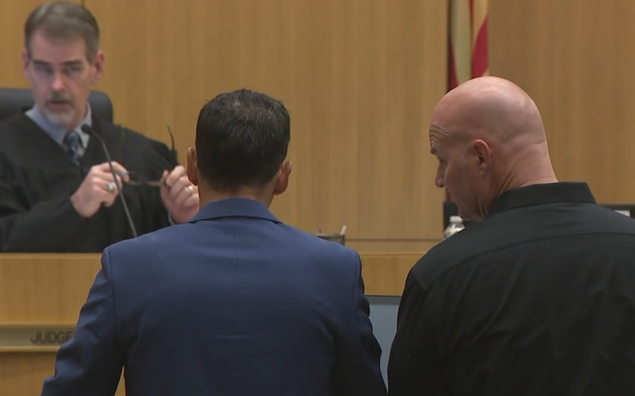 a man in black in a courtroom next to a man in a suit, both looking at a judge