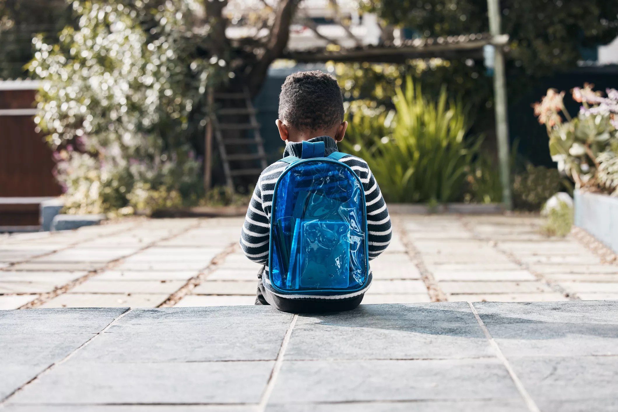 a sad young child with a backpack sitting, seen from behind