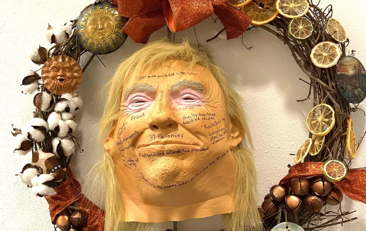 a wreath featuring a rubber trump mask with phrases like "defamation" and "sexual assault" written on its face