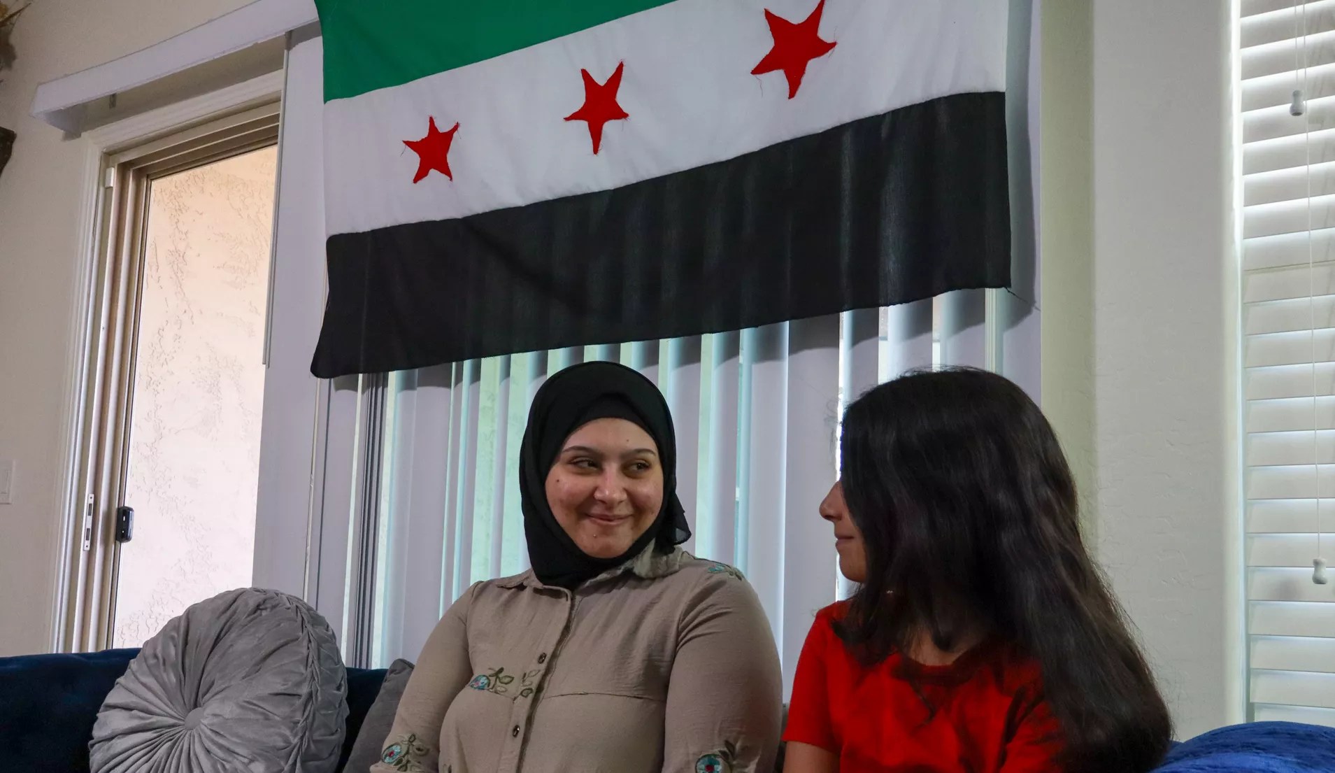 a syrian woman sits on a couch with an adolescent girl, a syrian flag hanging above them