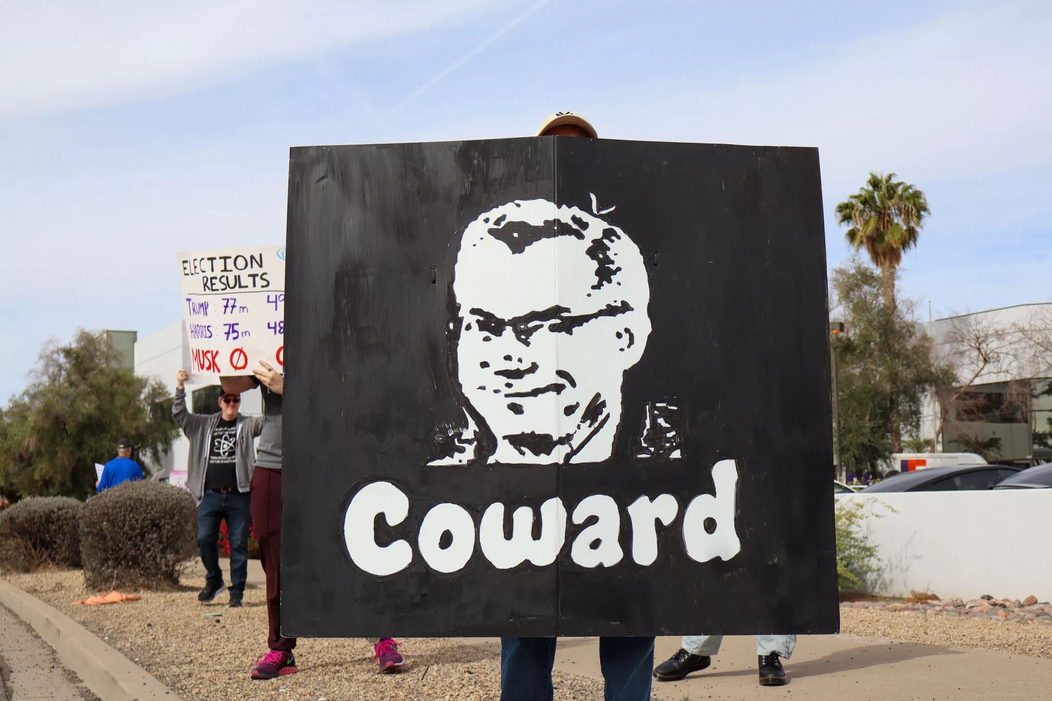 a protester holds a black sign with the face of rep. david schweikert and the word "coward"