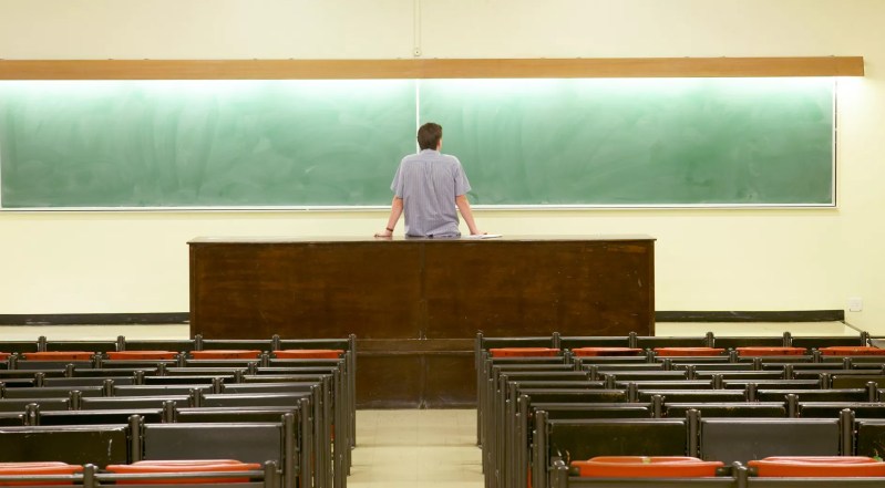 a stock photo of a professor staring at an empty chalkboard in an empty classroom