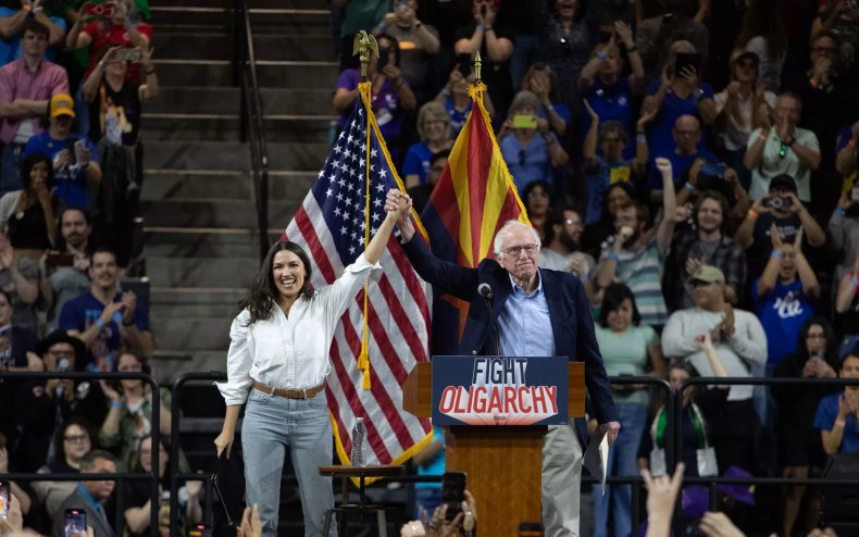 alexandria ocasio-cortez and bernie sanders hold hands high in the air in front of a U.S. flag and Arizona flag at a rally