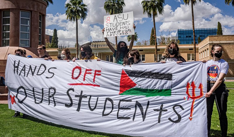 people hold a banner that says "hands off our students" with a Palestinian flag
