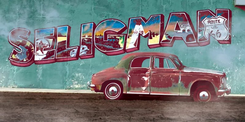 a rusted car in front of a wall with a postcard-style "Seligman" on the side