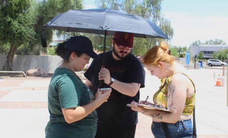 a woman watches another woman sign a form as a man holds an umbrella over both on a sunny day