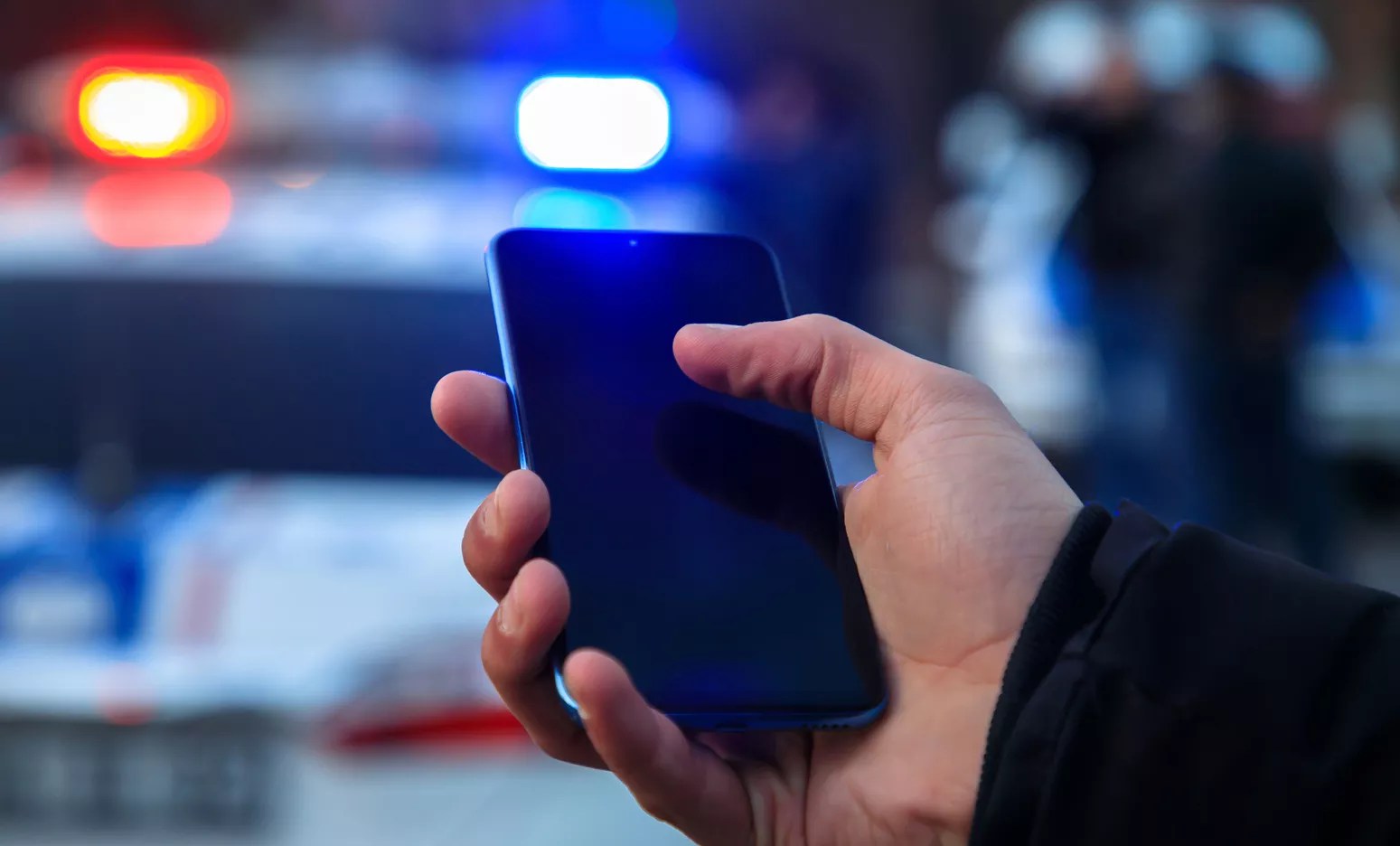 a hand holds a phone in front of a cop car with flashing lights